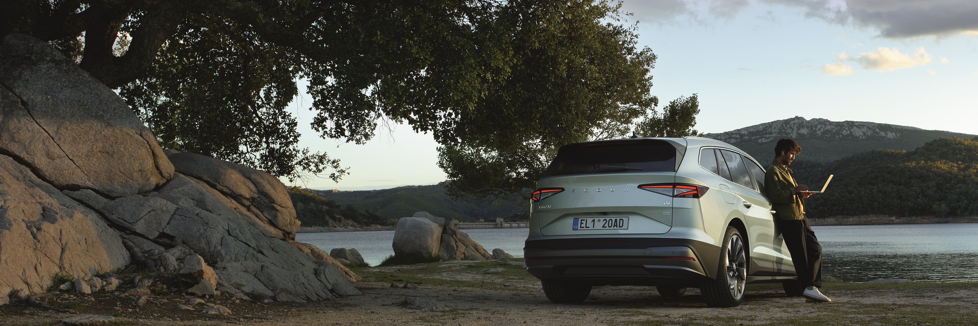 Ein Mann mit Laptop lehnt an einem Škoda Enyaq, dieser parkt an einem See